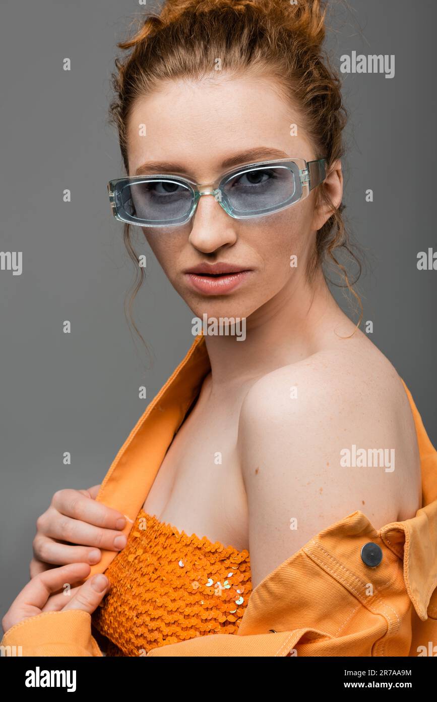 Confident young red haired woman in sunglasses posing in top with sequins and orange denim jacket while standing isolated on grey background, trendy s Stock Photo