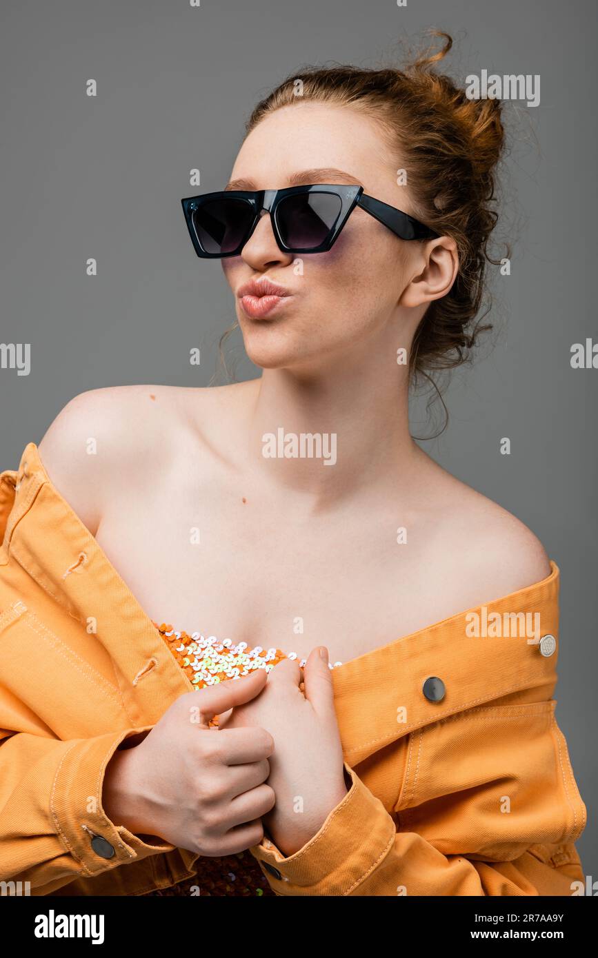 Portrait of young redhead woman in sunglasses, top with sequins and orange denim jacket pouting lips and posing isolated on grey background, trendy su Stock Photo
