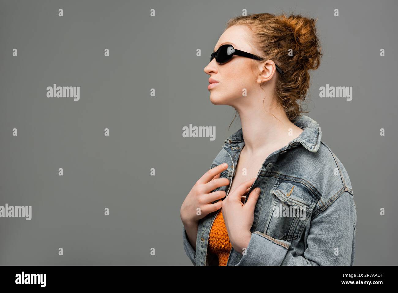 Young redhead model with natural makeup posing in sunglasses and top with sequins while touching denim jacket isolated on grey background, trendy sun Stock Photo