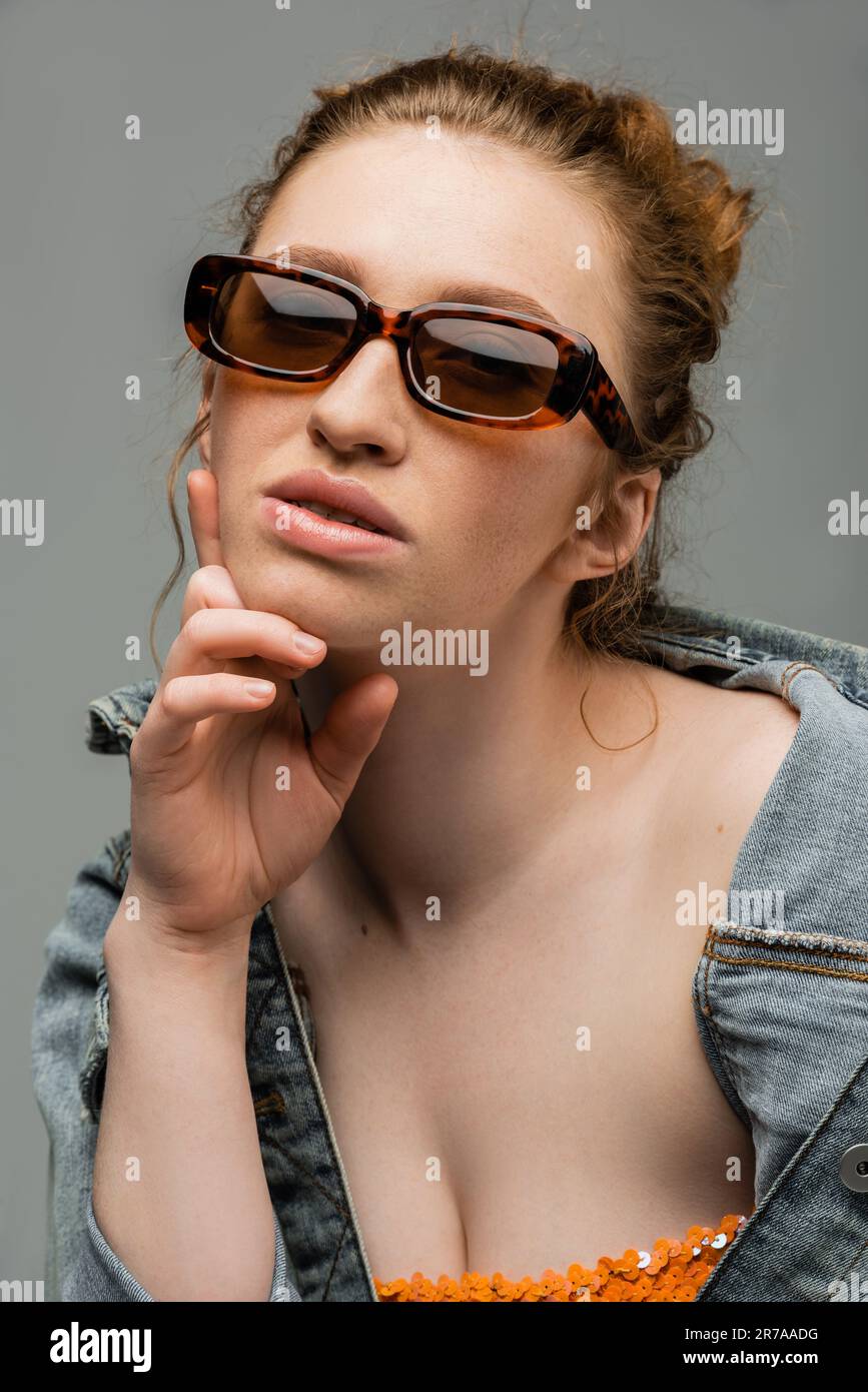 Portrait of young redhead woman in sunglasses posing in stylish denim jacket and top with sequins while standing isolated on grey background, trendy s Stock Photo