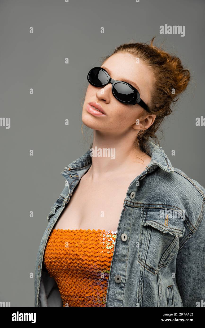 Confident and stylish young redhead woman in sunglasses, top with sequins and denim jacket standing and posing isolated on grey background, trendy sun Stock Photo