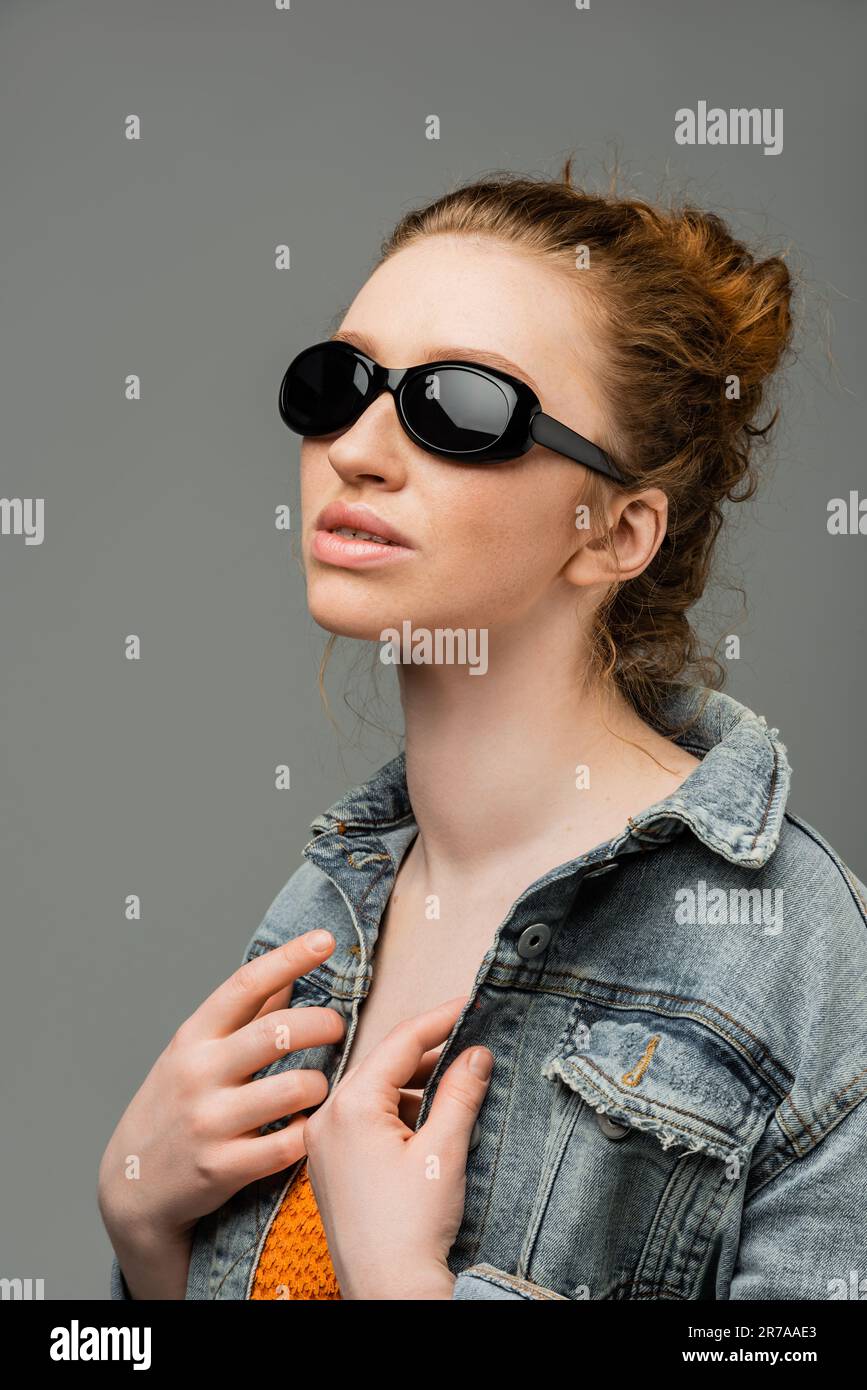 Red haired and freckled young woman in stylish sunglasses posing in denim jacket and top with sequins and standing isolated on grey background, trendy Stock Photo