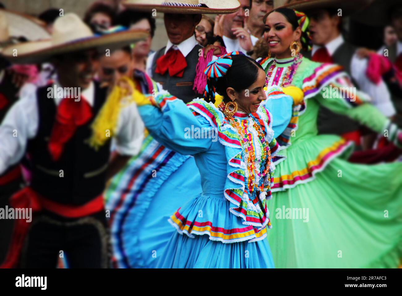 A group of men and women wearing traditional Mexican clothing, including colorful dresses and wide-brimmed hats, are dancing in a lively parade on a c Stock Photo