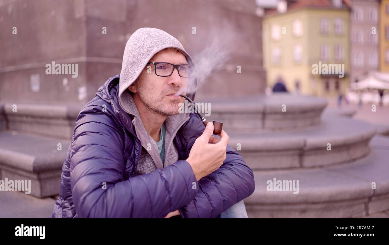 Adult man with glasses sitting on square and smoking a tobacco pipe releasing smoke in the Palace Square, Warsaw Old Town Stock Photo