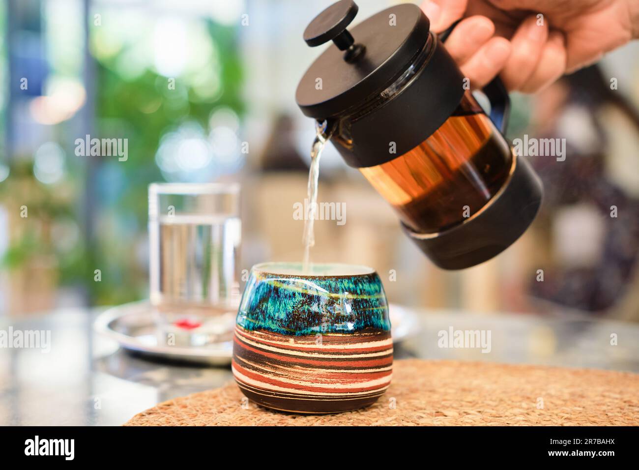 woman's hand pours tea from a French press into a beautiful handmade cup on a table in a cafe, selective focus, blurred background. Lifestyle, meeting Stock Photo