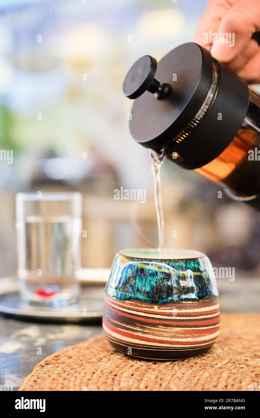 woman's hand pours tea from a french press into a beautiful handmade cup on table in a cafe, blurred background. Vertical frame, lifestyle meeting wit Stock Photo