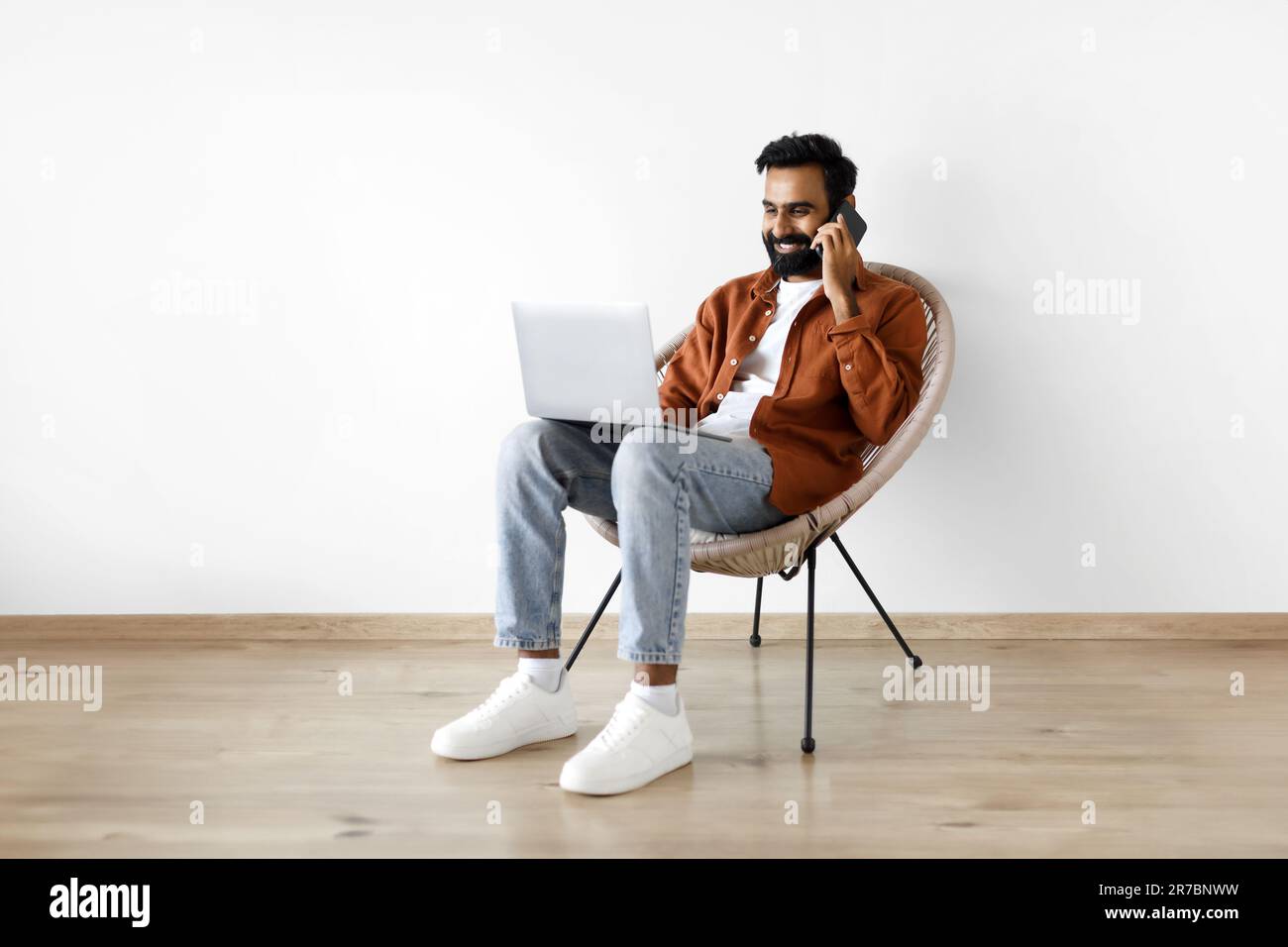 Indian Guy Talking On Cellphone Using Laptop Over White Wall Stock Photo