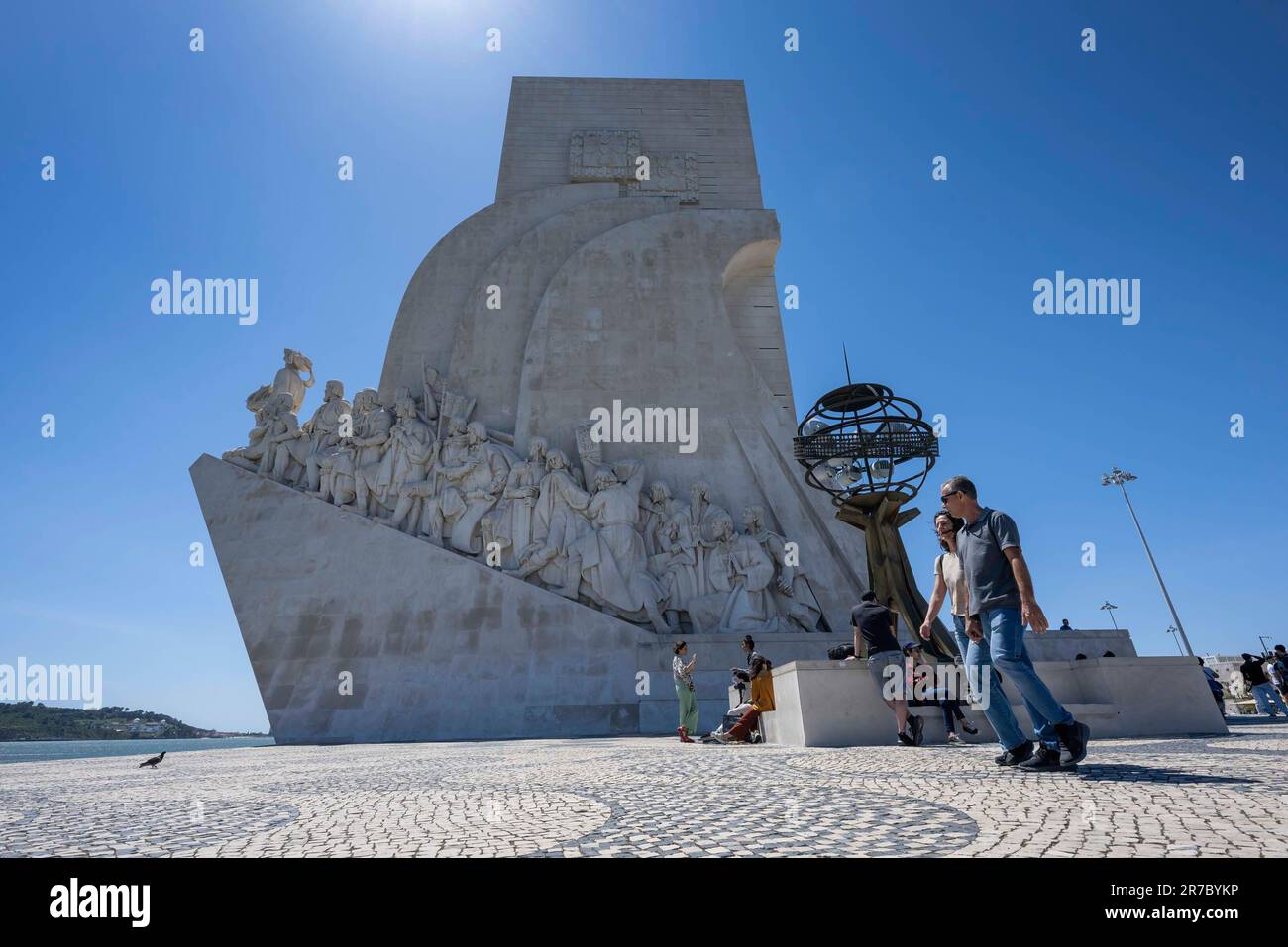 People are seen walking near the Monument to the Discoveries in the vicinity of the Rio Tejo promenade. The monument is a gigantic structure built in 1960 on the banks of the Tejo River in Belém, Lisbon, to commemorate the 500th anniversary of the death of Henry the Navigator and also to honor the memory of all those who were involved in the progress of the Age of Discoveries. Author of the work were the architect José Ângelo Cottinelli Telmo and the sculptor Leopoldo de Almeida, who were responsible for the sculptures. Stock Photo