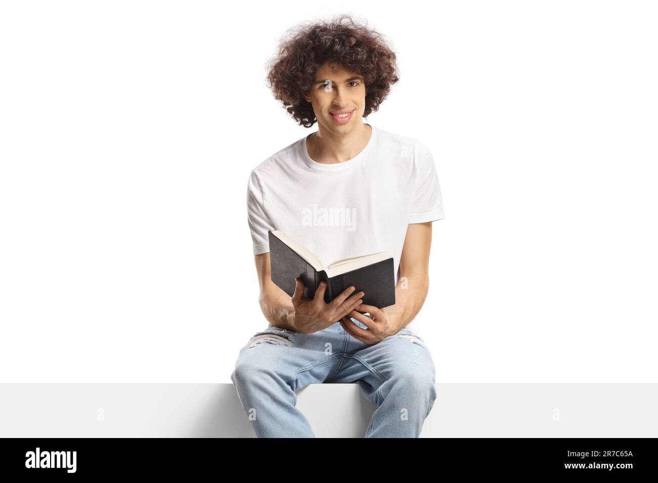 Young man with curly hair sitting on a panel and holding a book isolated on white background Stock Photo