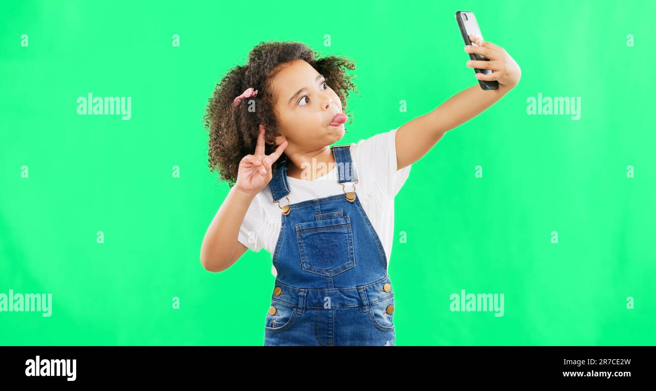 Silly little girl, selfie and peace sign on green screen with goofy facial expressions against a studio background. Female child or kid making funny Stock Photo