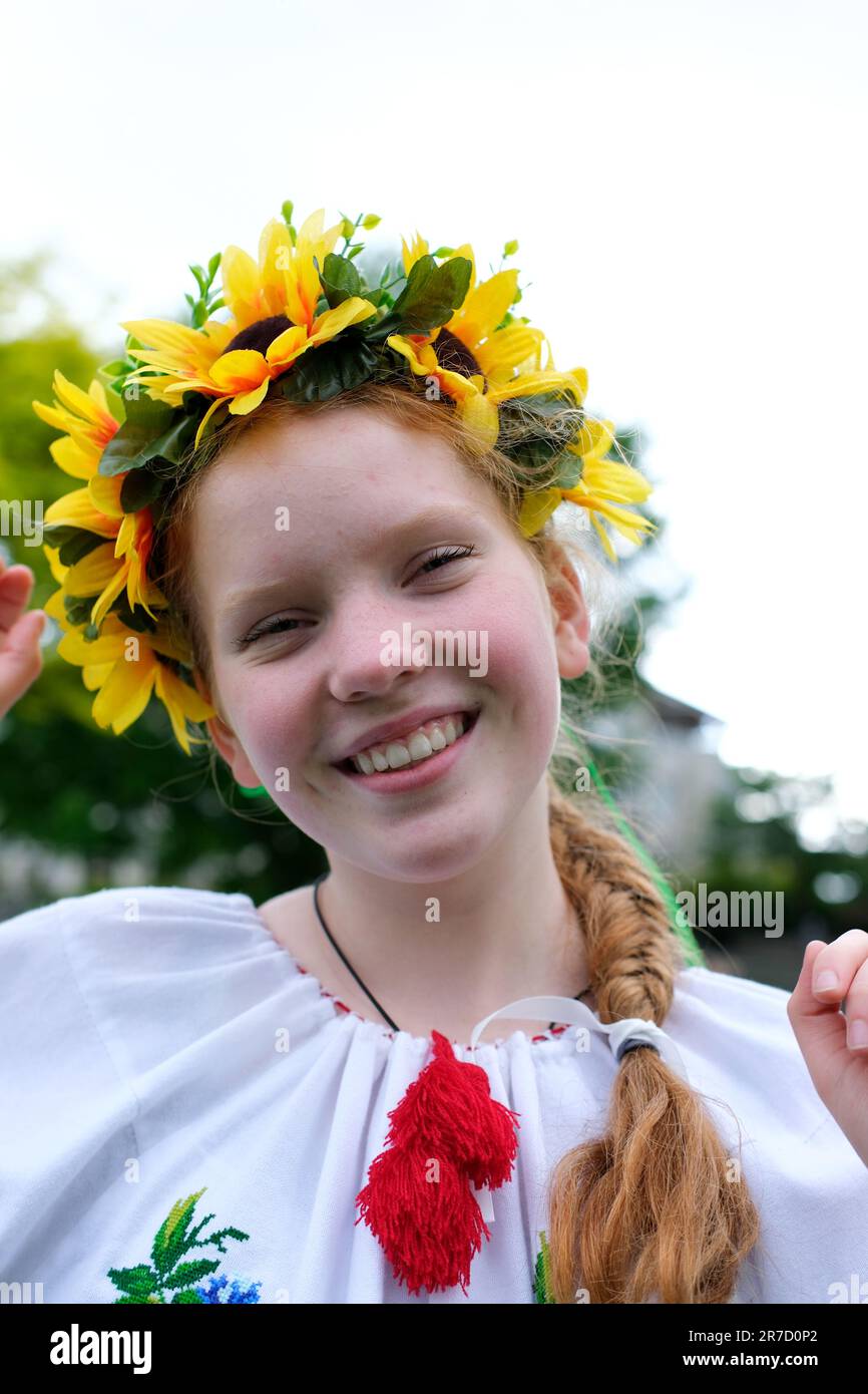 young red-haired girl in a wreath of sunflowers outdoors in park garden in beautiful embroidered Ukrainian vyshyvanka shirt with red braid with ribbons Ukraine Peaceful Time of happiness joy and peace Stock Photo