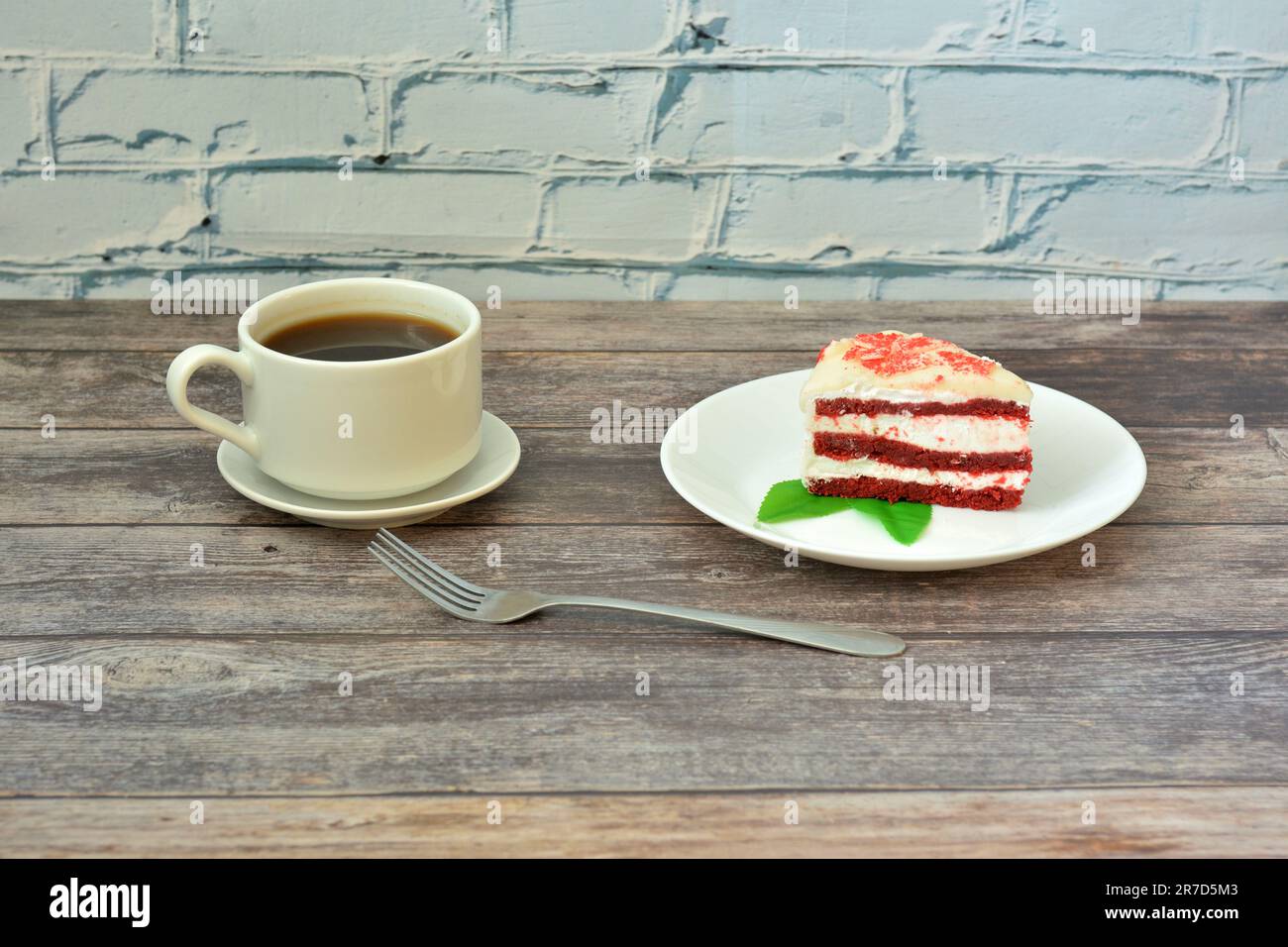 A cup of hot black coffee on a saucer and a plate with a piece of red velvet cake on a light wooden table. Close-up. Stock Photo