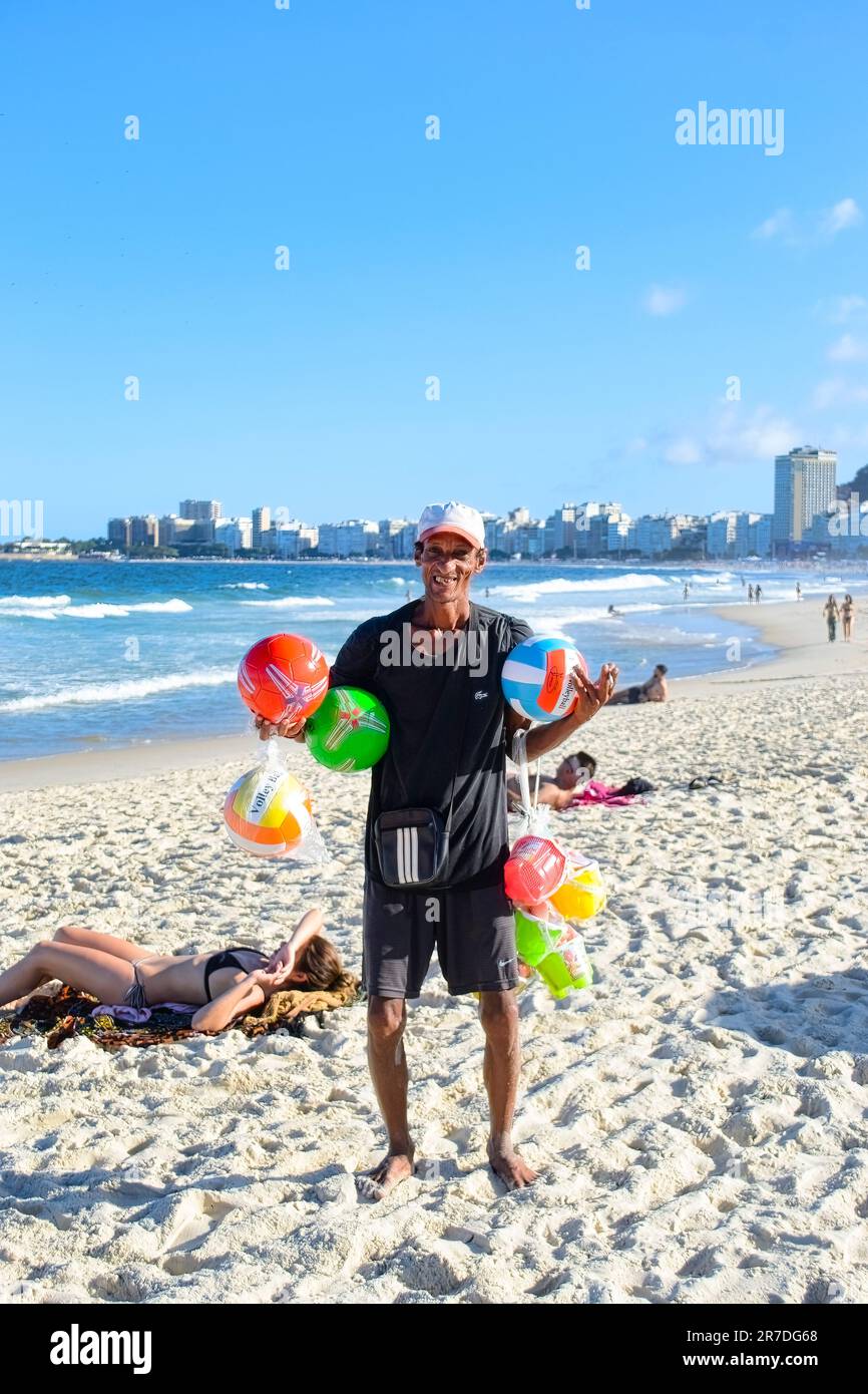 Rio de Janeiro, Brazil - May 25, 2023: A man stands, smiling and holding inflatable balls and other items on Copacabana beach. Two individuals sunbath Stock Photo