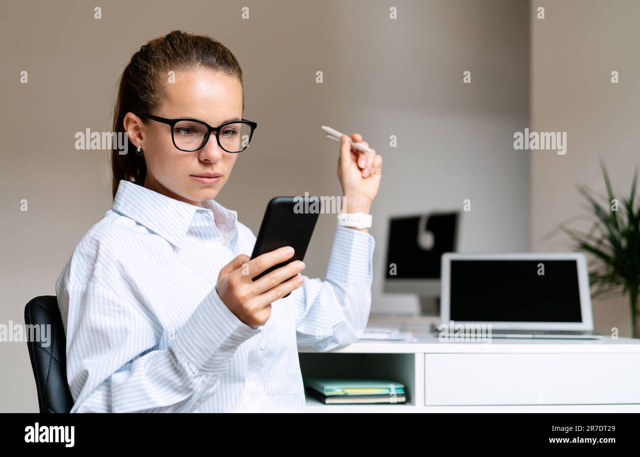 Serious young businesswoman wearing glasses sitting at the desk in her own office space and using her smart phone. Stock Photo