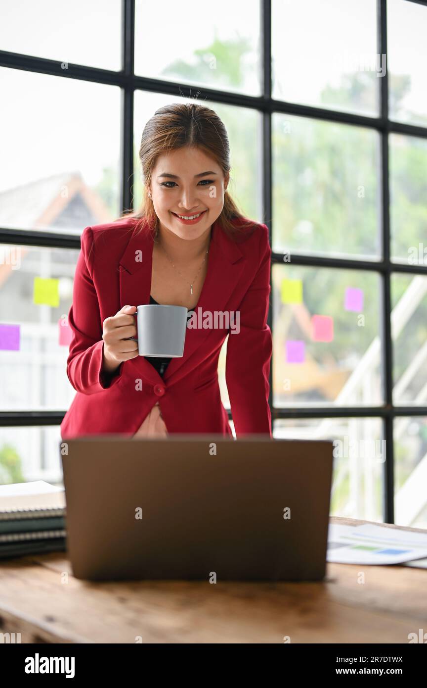 A portrait of a beautiful and attractive millennial Asian businesswoman in a red suit sipping coffee while working on her project on her laptop in the Stock Photo