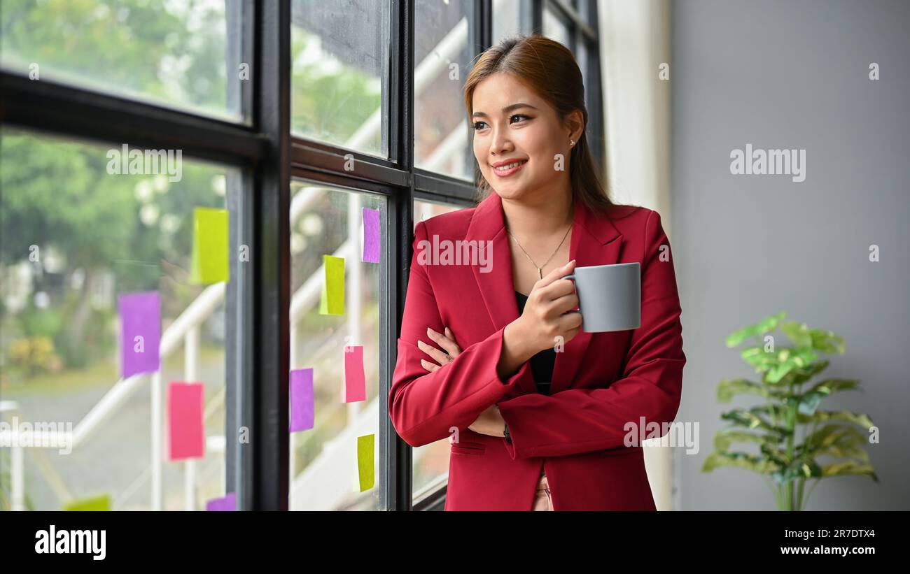 A portrait of a successful millennial Asian businesswoman or female CEO in a red suit stands by the window with a cup of coffee in her hand. Stock Photo