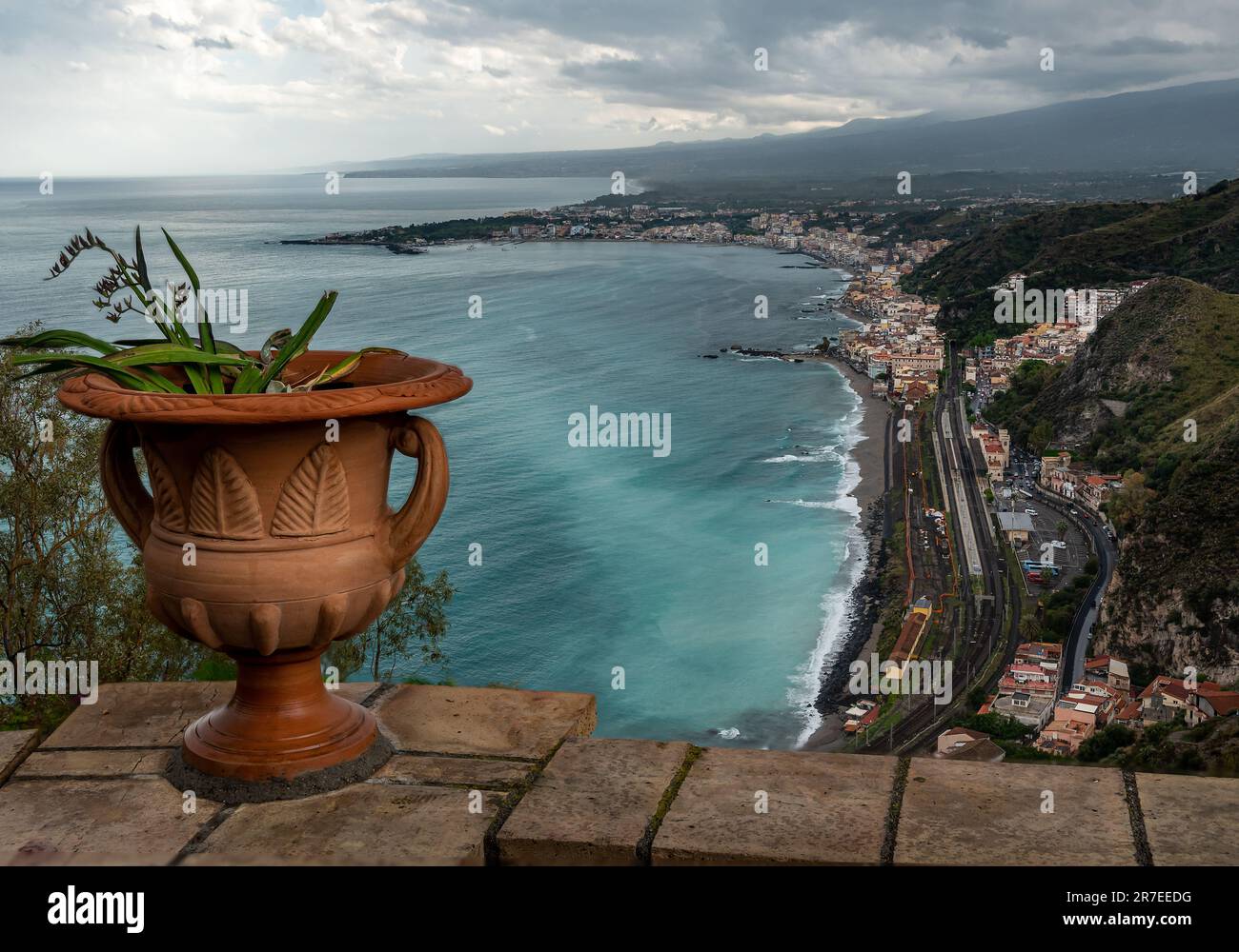 Mediterranean flowering plant in a stone ceramic pot with scenic view on seaside and Mount Etna volcano from public garden Parco Duca di Cesaro Stock Photo