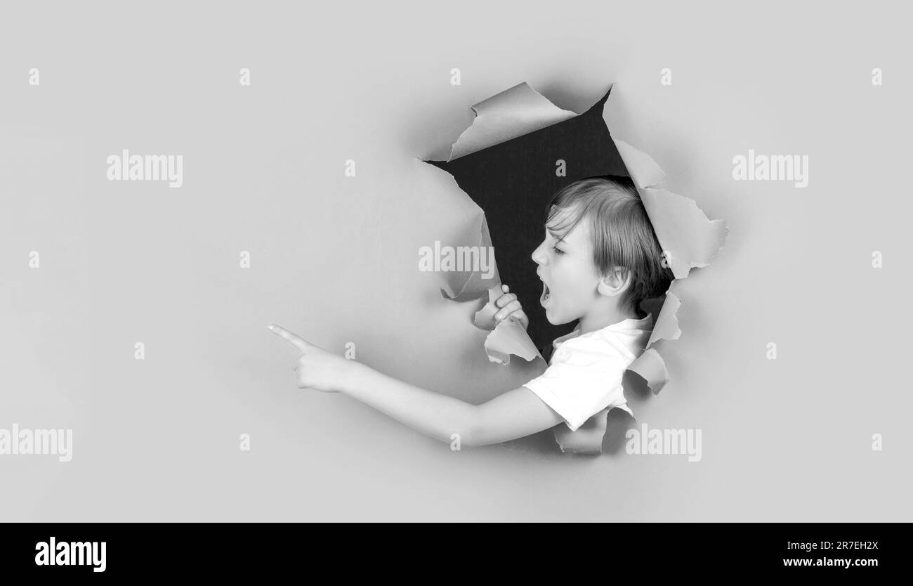 Screams boy looking through hole in paper wall. Shocked child making hole in paper. Kid looks through hole. Boy looks through ripped paper. Black and Stock Photo