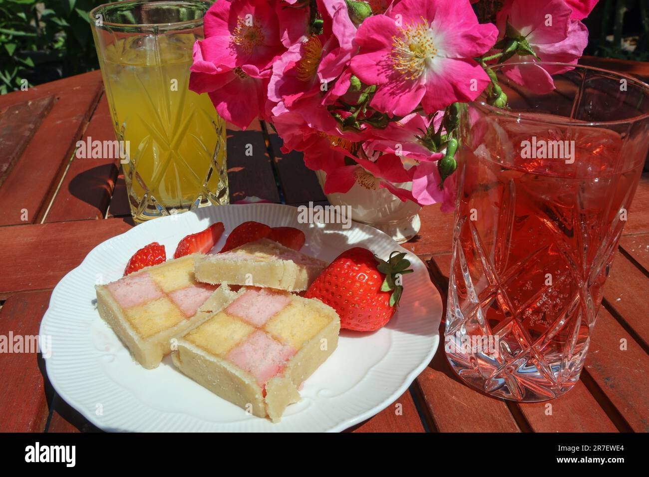 A plate of battenberg cake slices on a slated garden table with soft dronks and aposy of rambling roses; Stock Photo