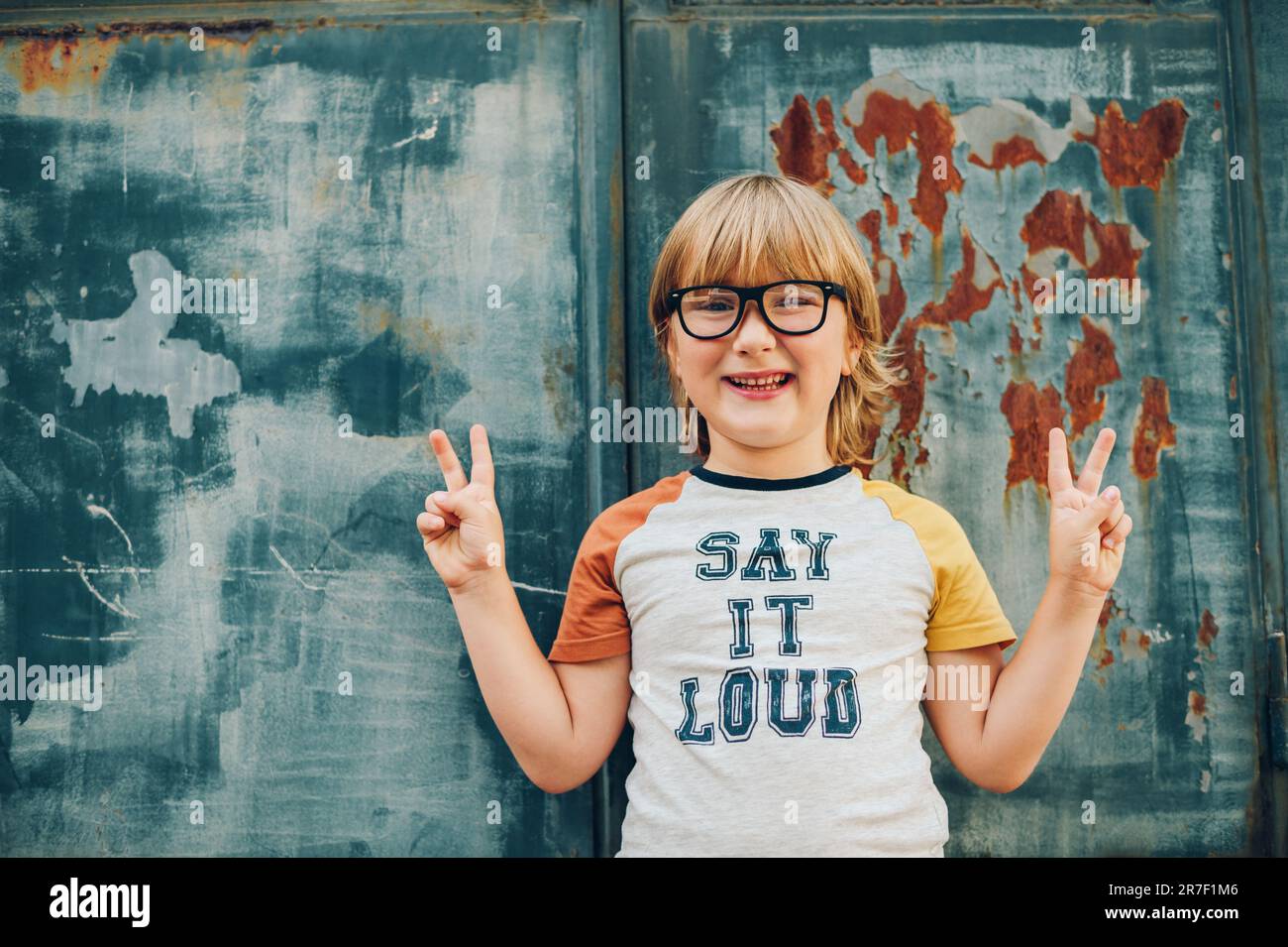 Outdoor portrait of funny little boy wearing glasses and t-shirt with sign 'Say It Loud' Stock Photo