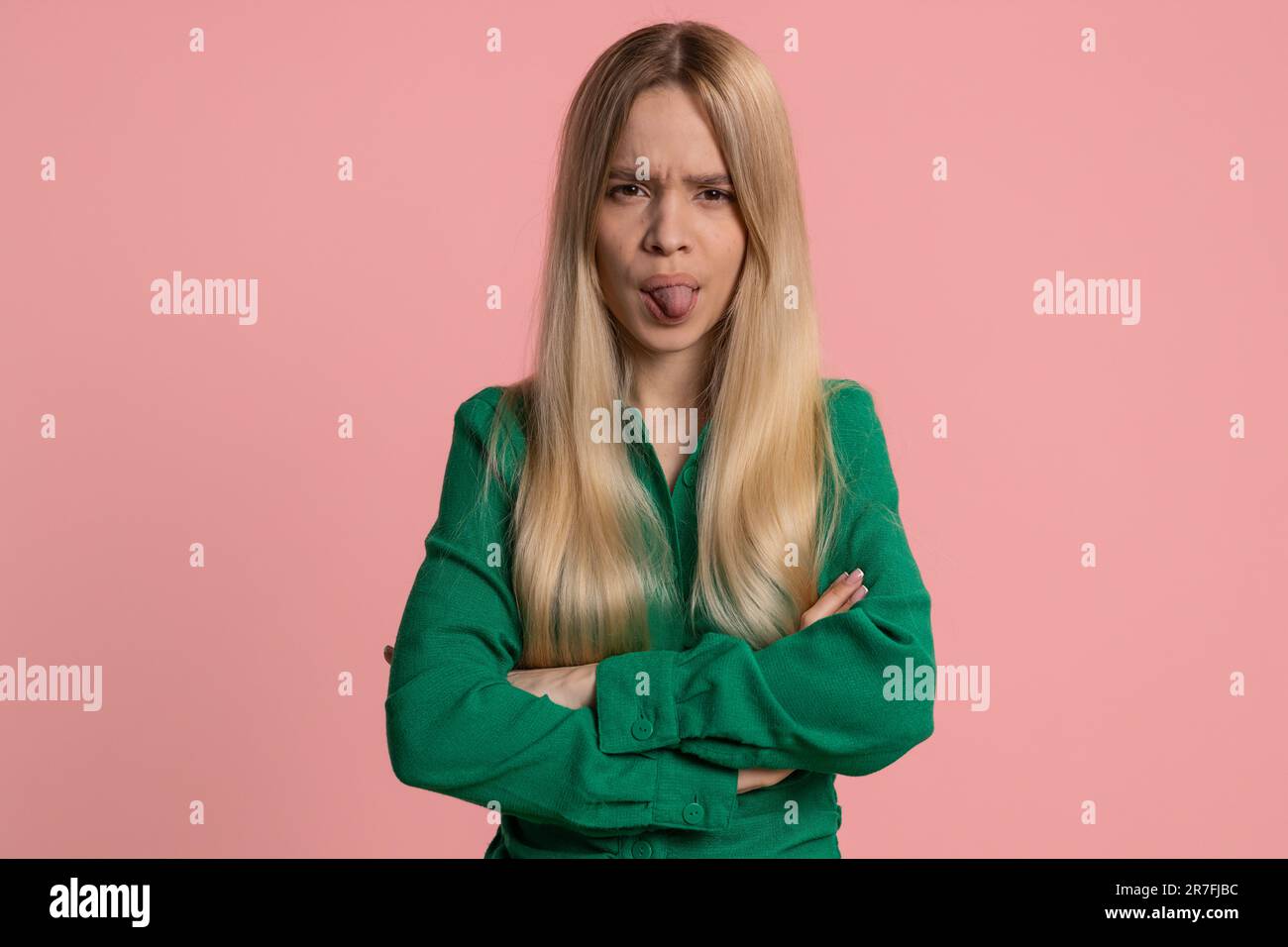 Young caucasian woman showing tongue making childish faces at camera, fooling around, joking, aping with silly face, teasing, bullying abuse. Pretty girl isolated on studio pink background indoors Stock Photo