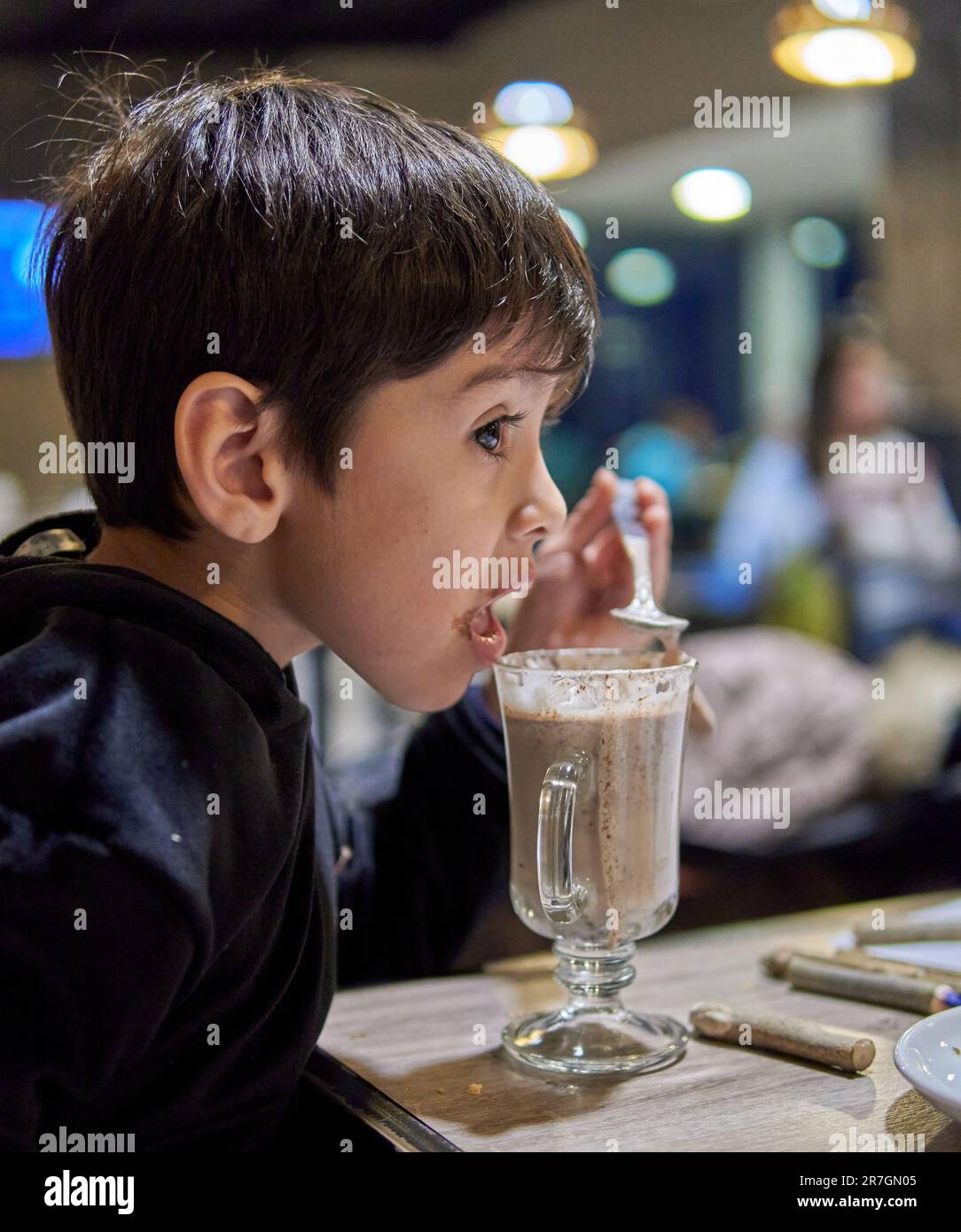 Brown little boy in profile in a bar drinking hot chocolate milk with a spoon. Out of focus background Stock Photo