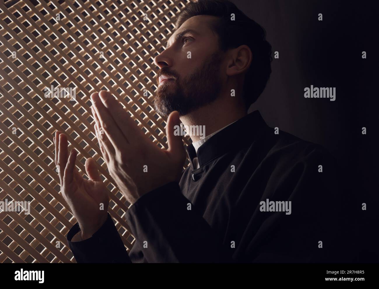 Catholic priest in cassock praying to God in confessional booth Stock Photo