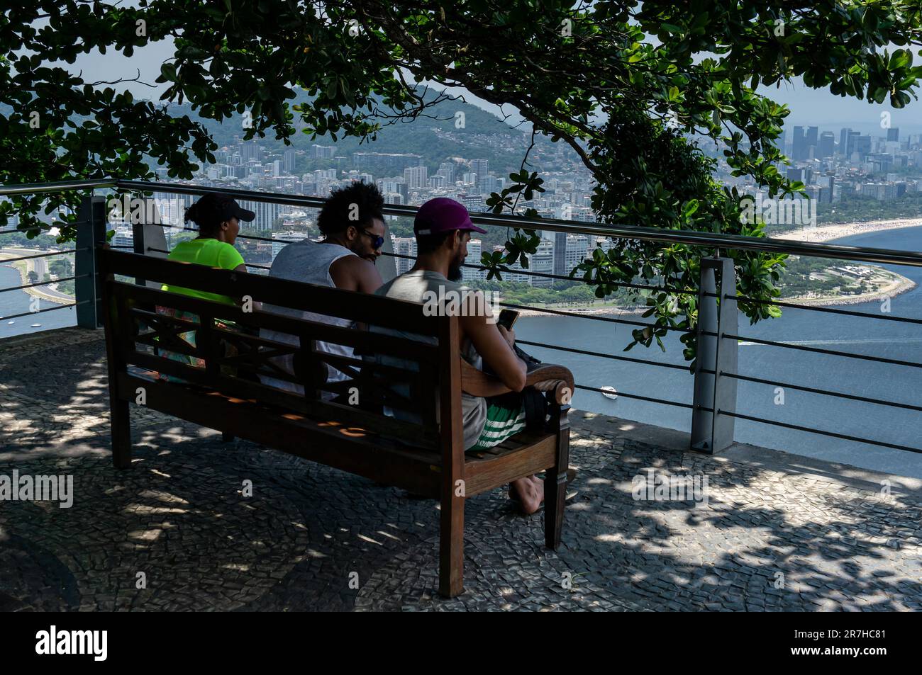 Tourists taking some time seated on a wood bench under tree shade at the observation deck guard rail of Urca hill in a summer afternoon sunny day. Stock Photo