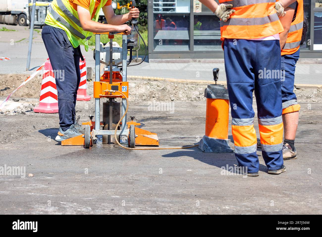 Road workers in reflective capes drill asphalt with a core drilling machine to take and measure cores during road repairs on a sunny day. Copy space. Stock Photo