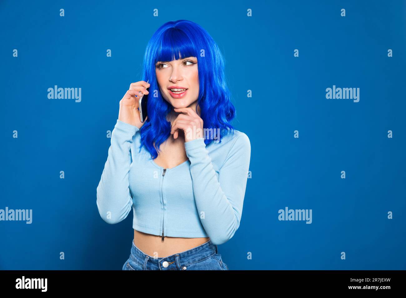 Confident young female model with bright dyed hair in casual clothes smiling and looking away while talking on smartphone against blue background Stock Photo