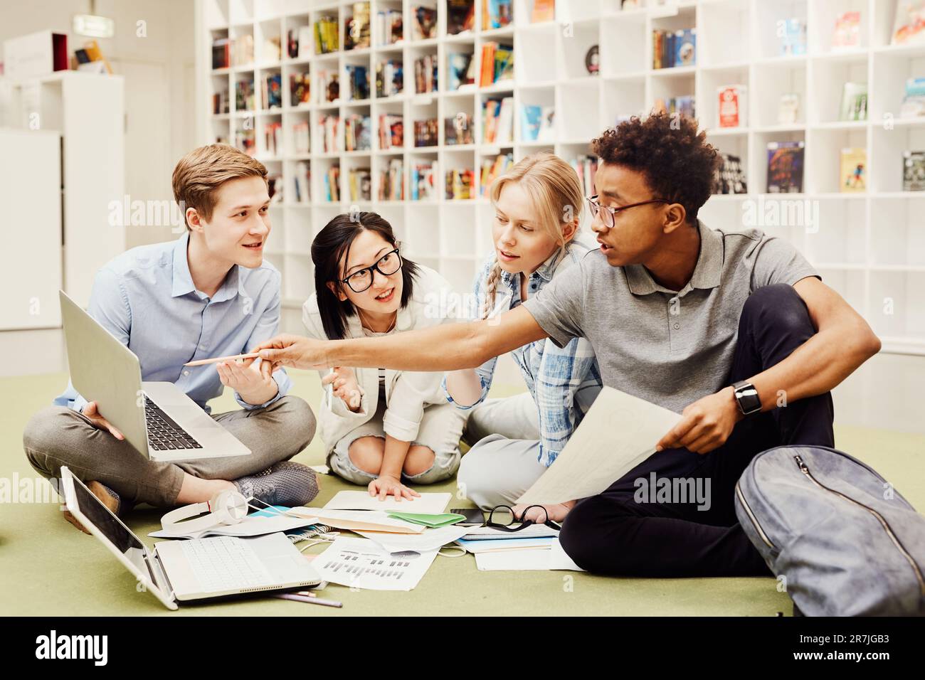 Serious confident young black guy sitting on floor in library and pointing at laptop screen while explaining information to groupmates Stock Photo