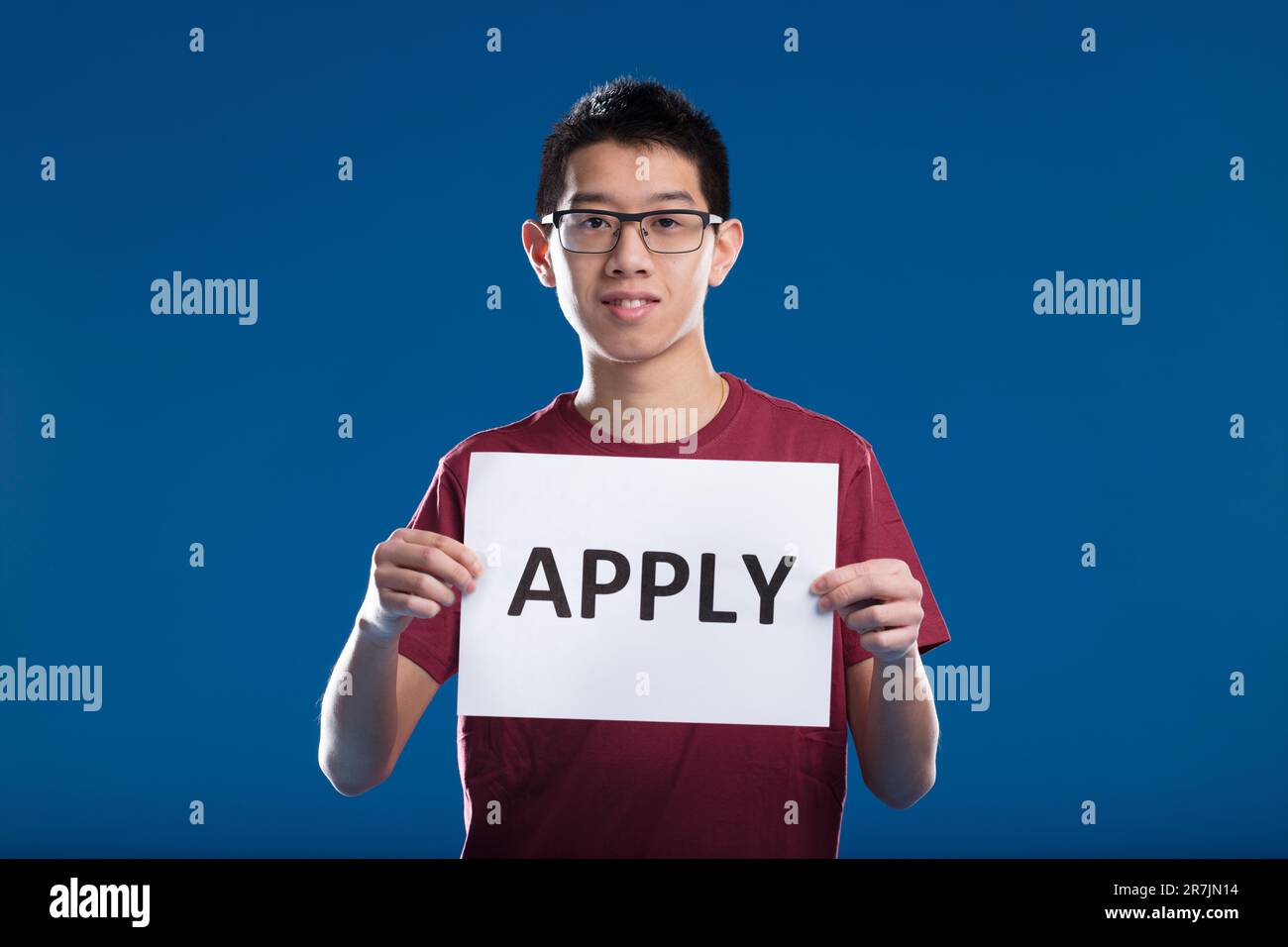 Young Asian man holding a sign saying APPLY. Inviting you to work in STEM at his company, wearing glasses and a burgundy shirt on a blue backdrop Stock Photo