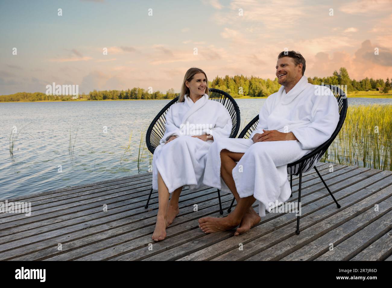 happy couple in white bathrobes relaxing on wooden lake footbridge after spa treatments at warm summer evening Stock Photo