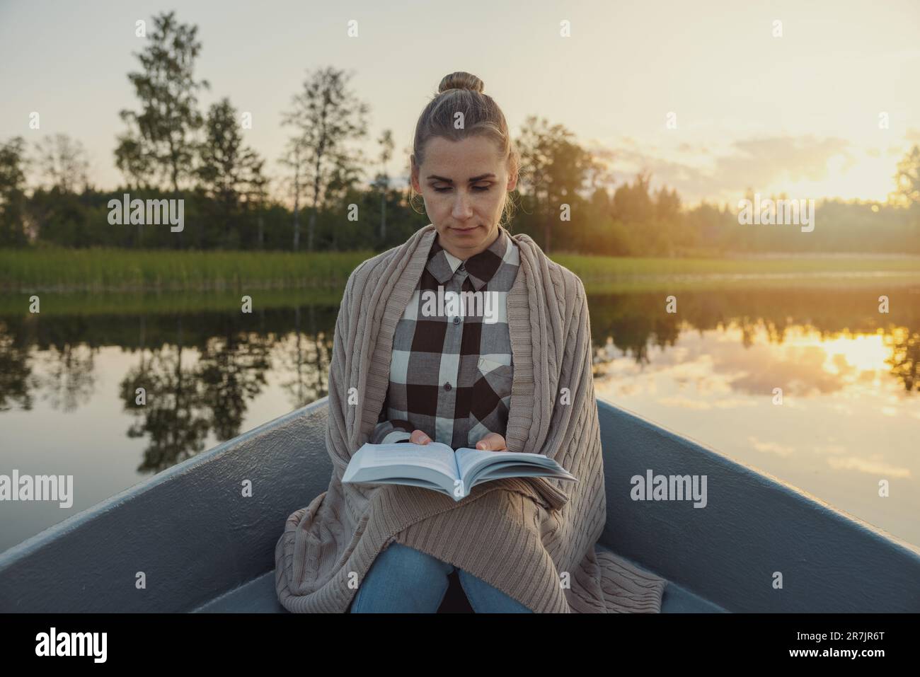 woman with blanket on shoulders sitting in a row boat on the lake and read a book at sunset Stock Photo