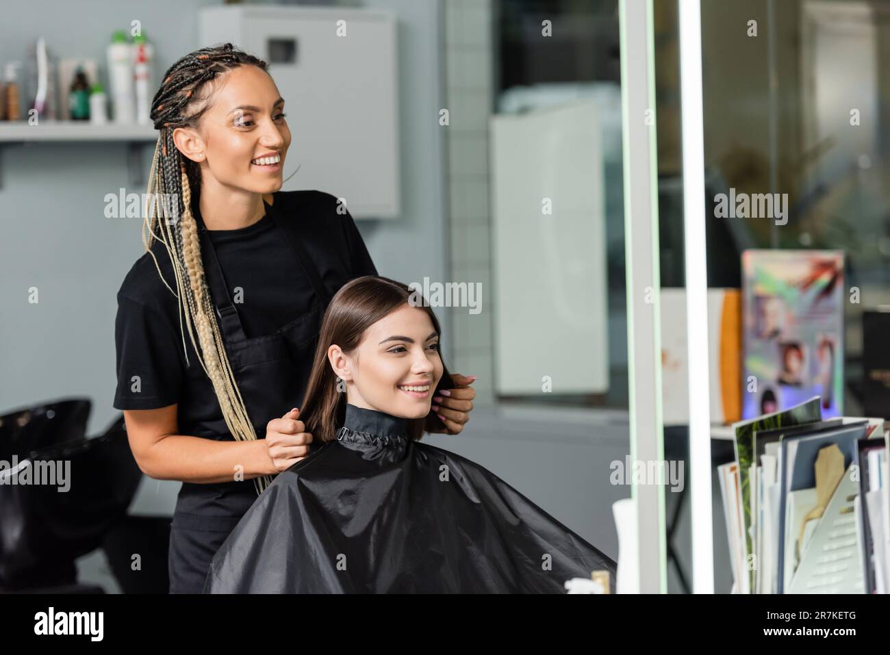 beauty industry, positivity, happy hair stylist with braids wearing hairdressing cape on female client, hair extension, hair treatment, salon customer Stock Photo