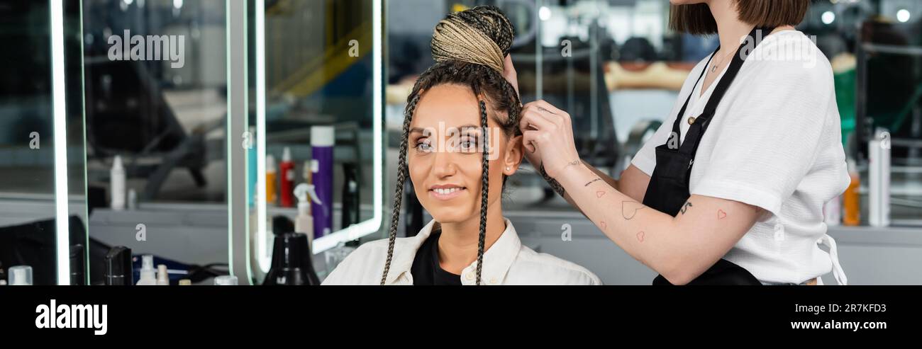 salon experience, tattooed hairdresser doing hair bun to female client with braids, cheerful women, client satisfaction, customer in salon, beauty ser Stock Photo