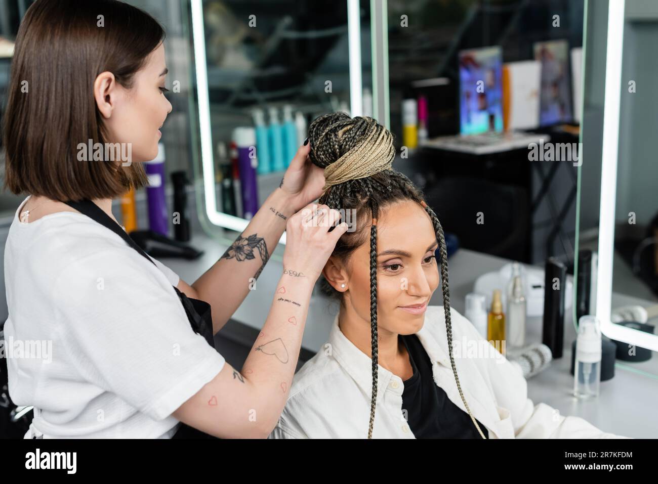 salon experience, tattooed hairdresser doing hair bun to female client with braids, happy women, client satisfaction, customer in salon, beauty servic Stock Photo
