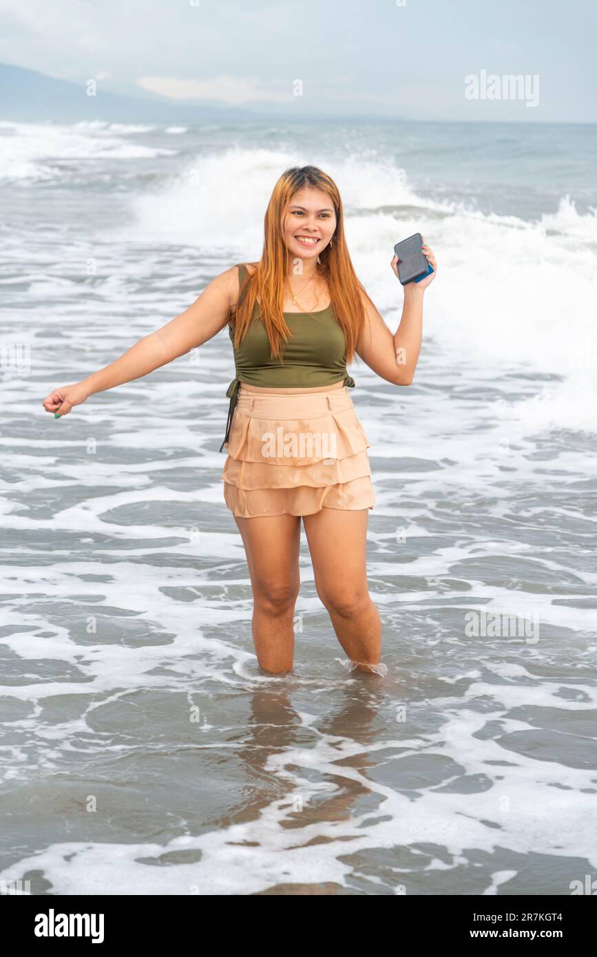 Holding her phone for selfies,on a beach near Manila at dusk,aimlessly strolling the warm shoreline waters,along smooth sands,having fun,dancing aroun Stock Photo