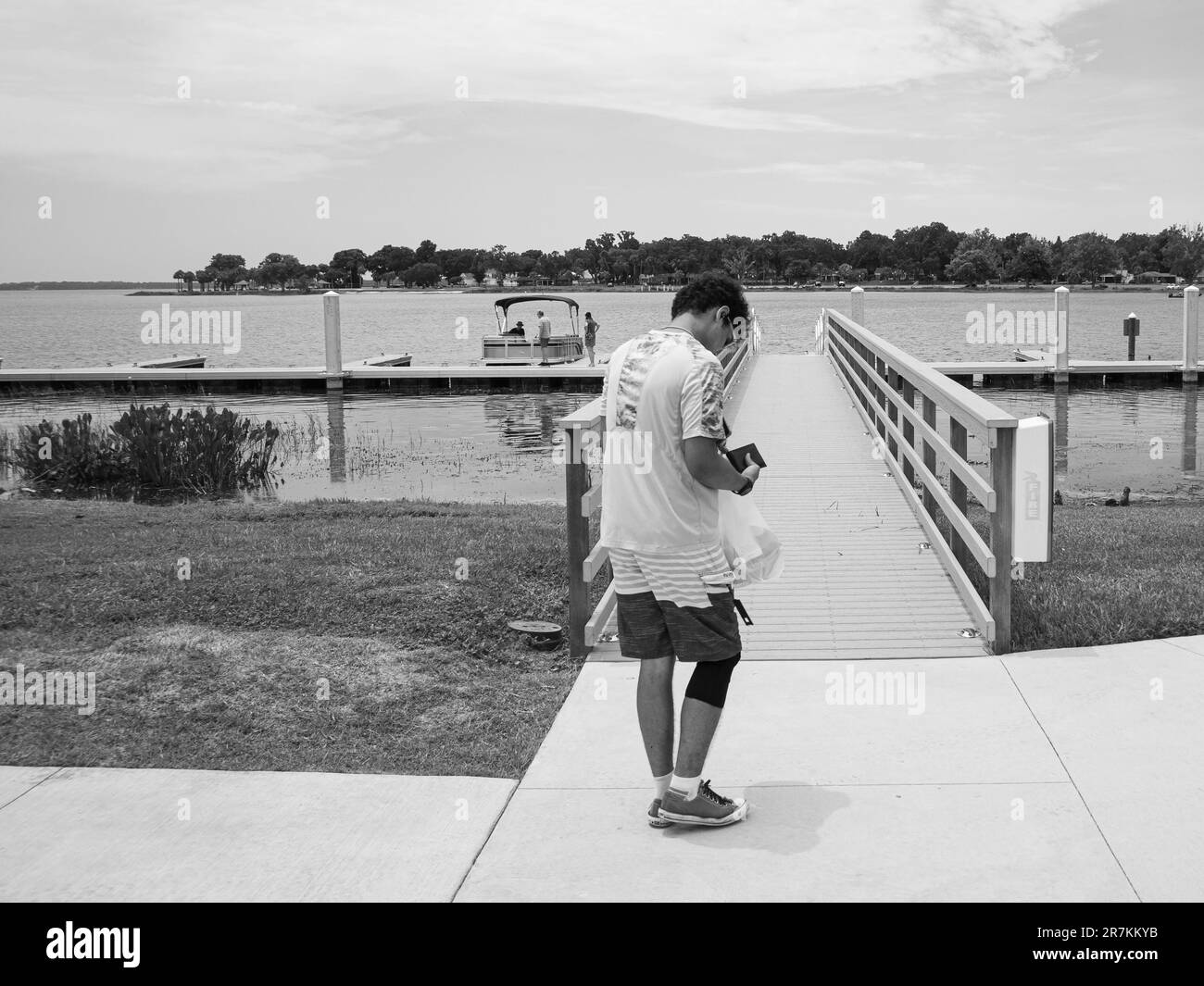 Black and white photo of young man standing near bridge at lake checking his phone. Stock Photo