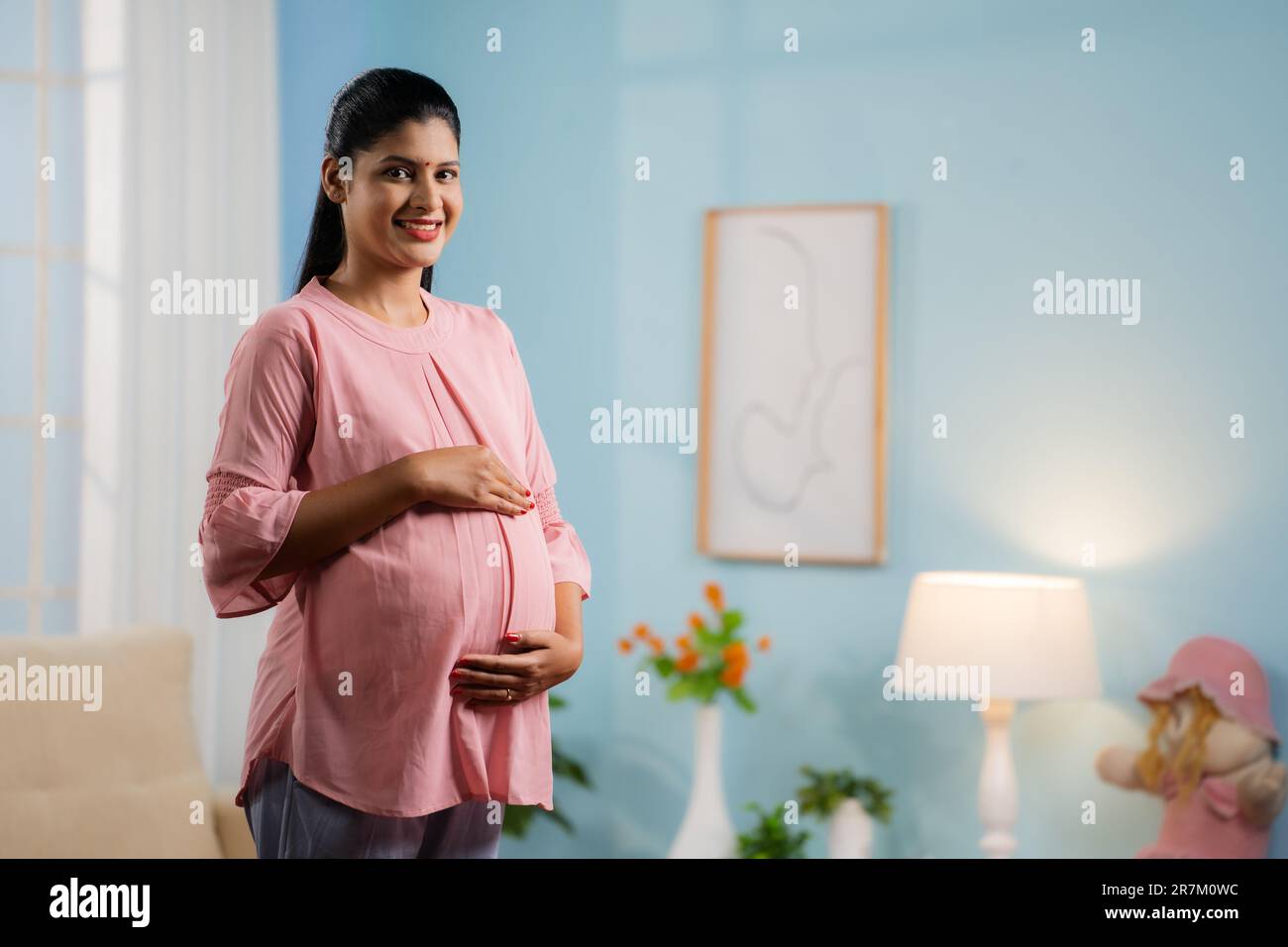 Happy smiling Indian Pregnant woman standing by feeling tummy while looking camera at home - concept of motherhood, expecting baby and parenthood Stock Photo