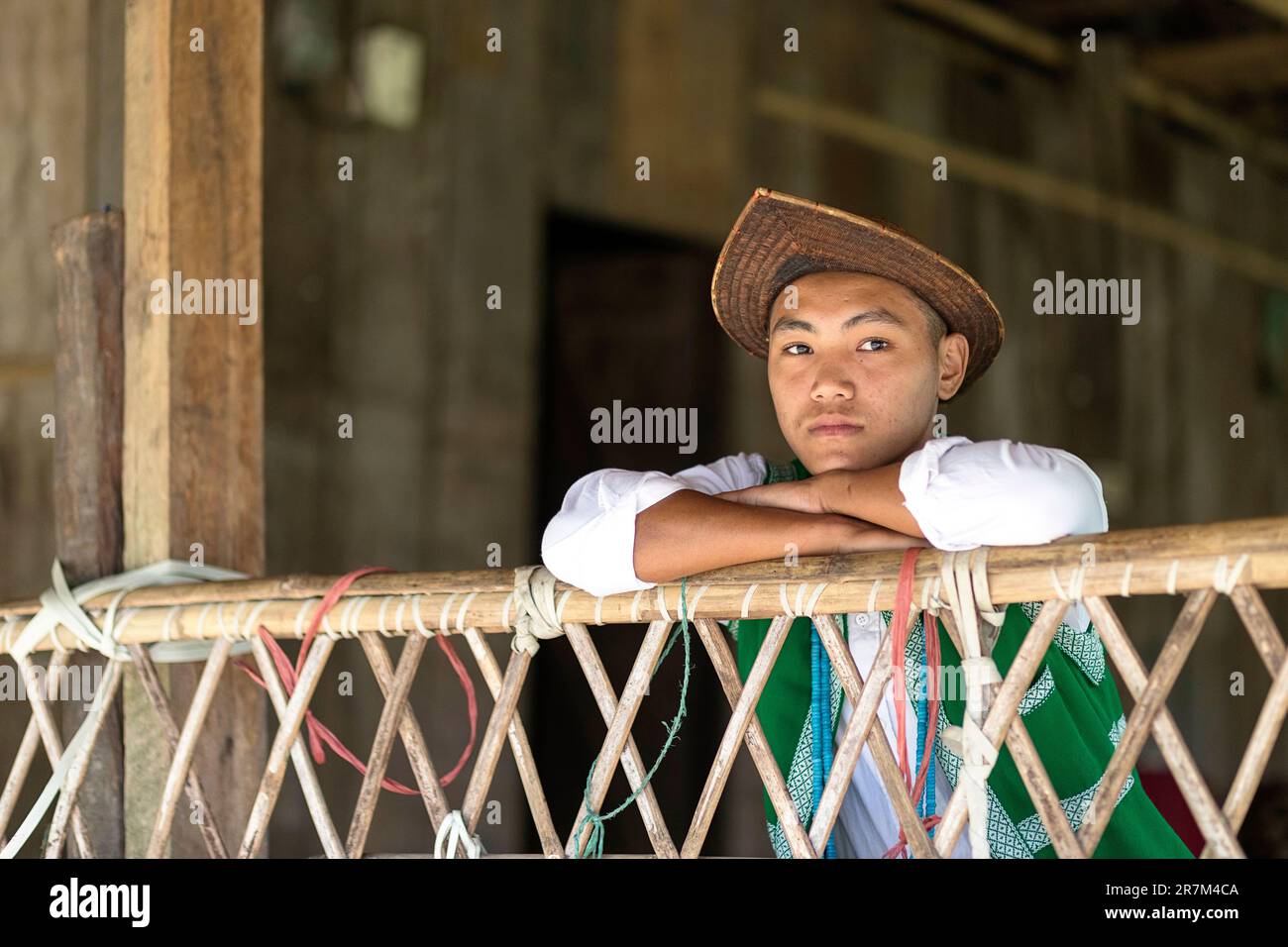 Boy from galo tribe wearing traditional cane hat and traditional clothes leaning his head on the arms on the bamboo fence at his house, Assam, India Stock Photo