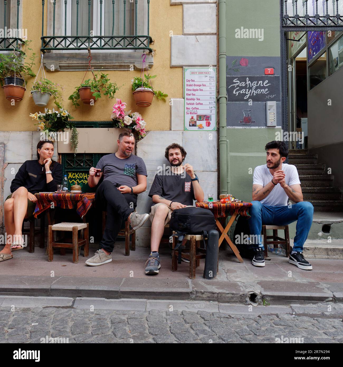 Friends enjoying a Turkish tea on informal table and chairs on a pavement in a quaint street with flower pots in the Galata area of Istanbul, Turkey Stock Photo
