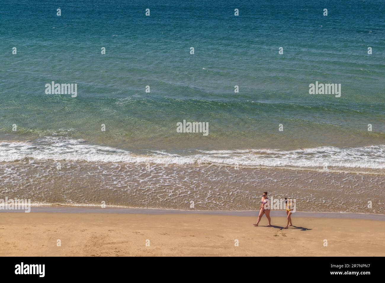 Two young Caucasian girls walking in bikinis along the sand, on the shore of the Mediterranean Sea in the town of Salou, Tarragona, Catalonia, Spain. Stock Photo