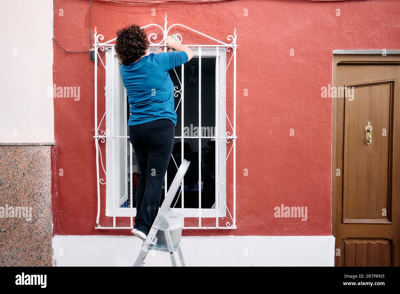 An elderly curly-haired woman on an aluminum ladder is cleaning the ironwork on the window of her home. Stock Photo