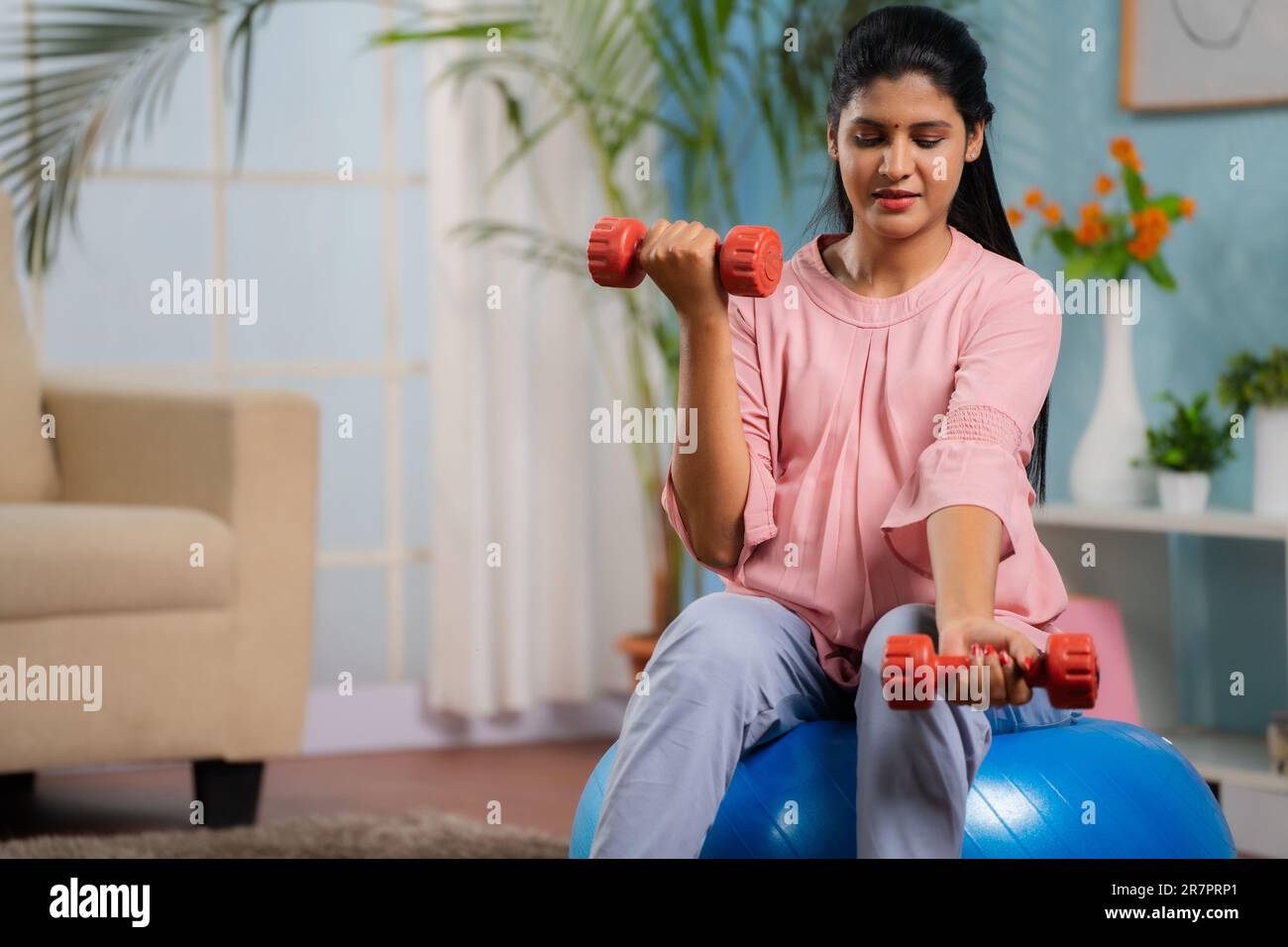 Indian pregnant woman doing exercise using dumbells while sitting on fitness ball at home - concept of active pregnancy, healthy lifestyle and Stock Photo