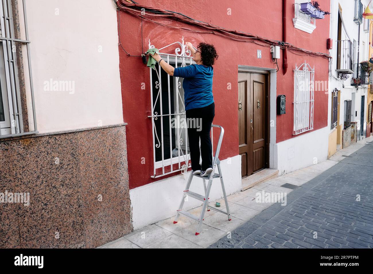 An elderly curly-haired woman on an aluminum ladder is cleaning the ironwork on the window of her home. Stock Photo