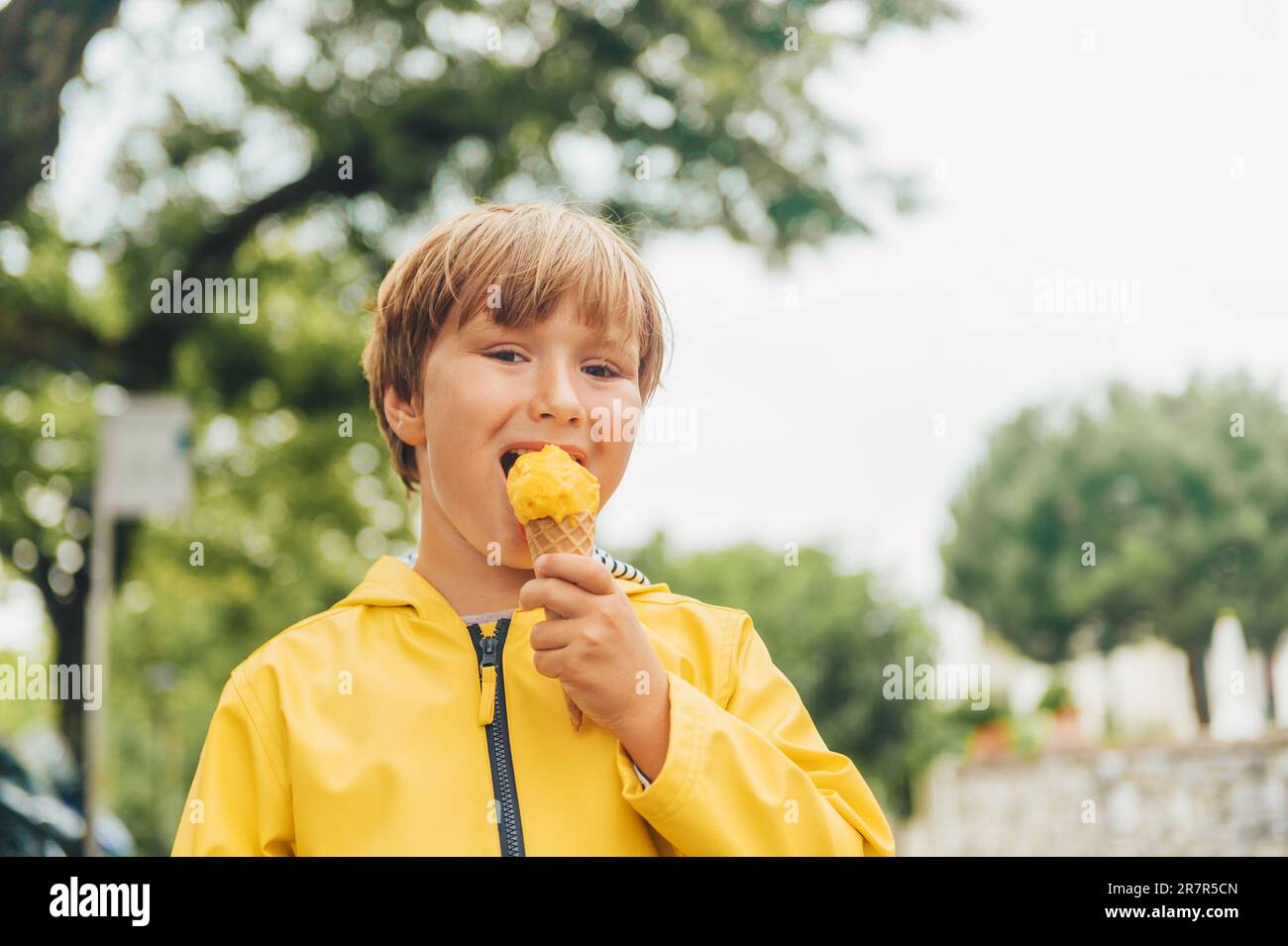 Cute little boy wearing yellow rain coat, eating mango ice cream Stock