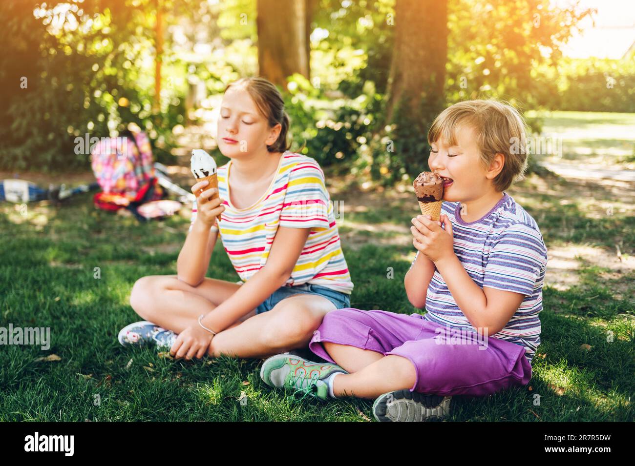 Two funny kids eating ice cream in summer park, sitting on the grass, wearing colorful stripe t-shirt Stock Photo