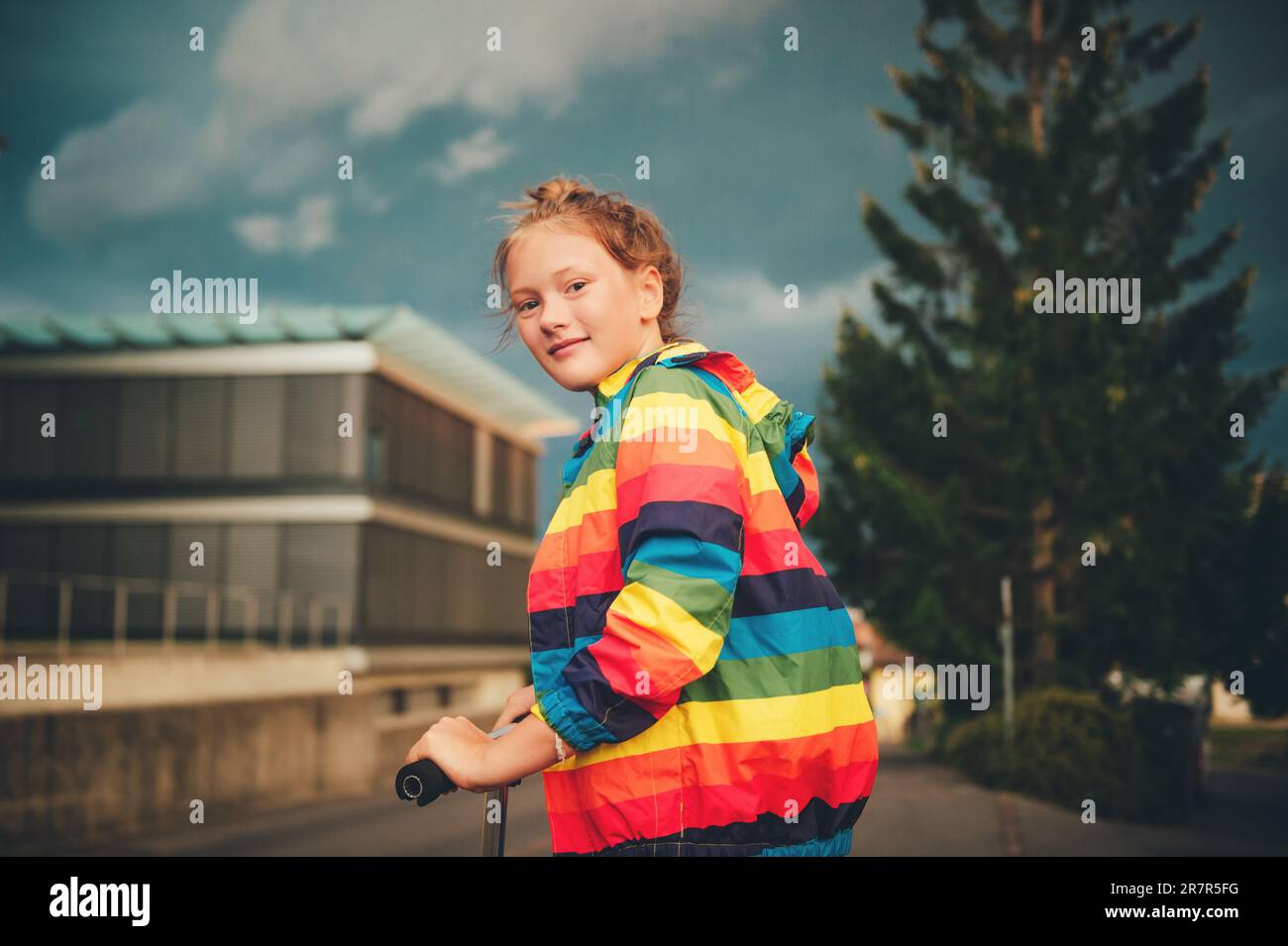 Young girl riding scooter outside in the evening, wearing colorful jacket Stock Photo