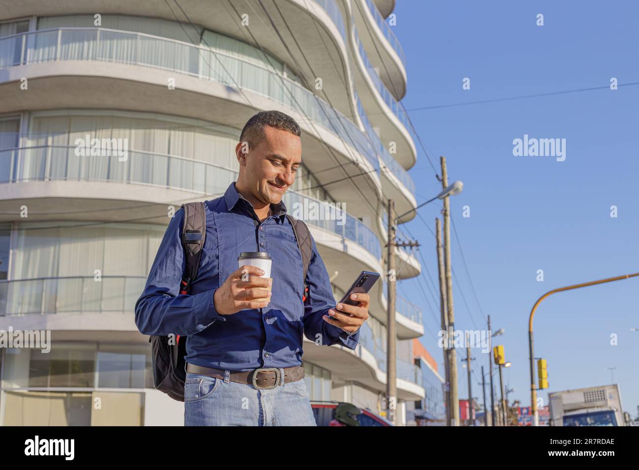 Young latin man looking at his mobile phone and holding a paper cup with coffee in his hand with copy space. Stock Photo